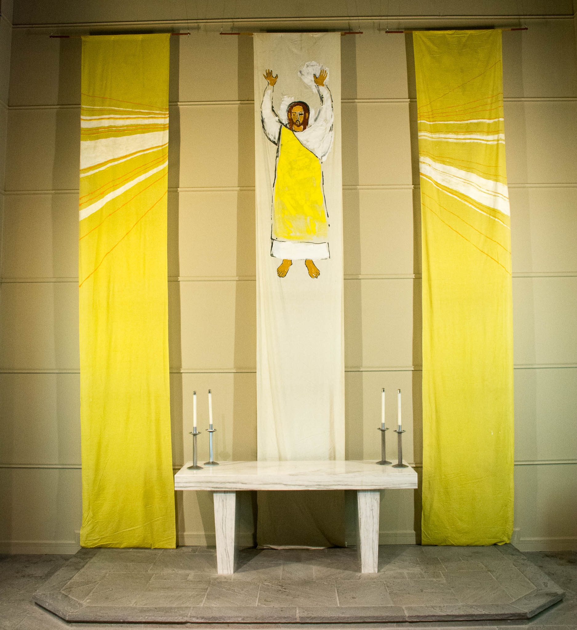 Three vertical banners hang behind the altar. In the middle, Jesus reaches into the air. Bright yellow panels with white rays of light hang on either side.