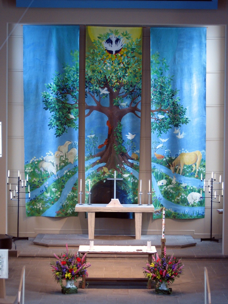 Photograph of three large cloth paintings behind an altar, depicting a large tree, with animals and birds around it, against a blue sky.