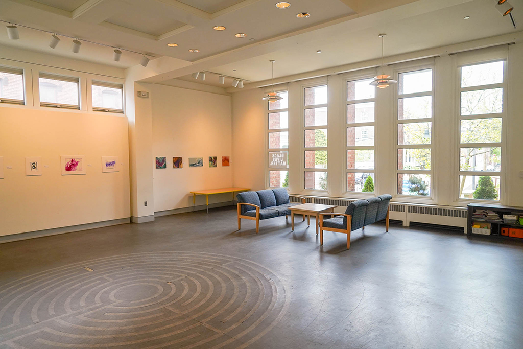 The Alumni/ae Room showing an art exhibit of brightly colored paintings on the wall, and light coming in the full-length windows.