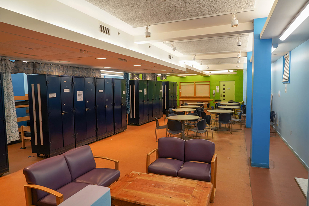 A large, well-lit room includes comfortable chairs around a coffee table, lockers, and round eating tables with chairs.