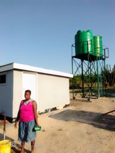 A woman waits outside for water. Two green water tanks are in the background.